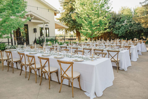Outdoor wedding reception venue with long tables set with white linens and wooden chairs under string lights.