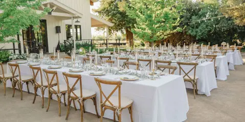 Outdoor wedding reception venue with long tables set with white linens and wooden chairs under string lights.