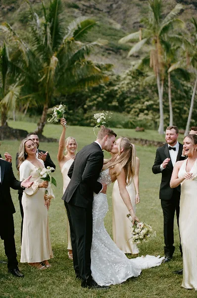A bride and groom share a kiss on a grassy lawn surrounded by their wedding party, with palm trees and a hillside in the background.