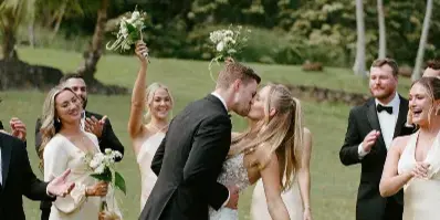 A bride and groom share a kiss on a grassy lawn surrounded by their wedding party, with palm trees and a hillside in the background.