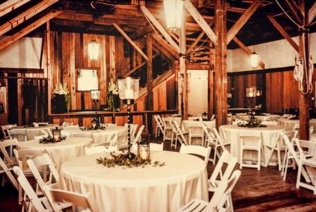A rustic wooden event hall set up for a wedding with round tables covered in white linens, white Chiavari chairs, and floral centerpieces.