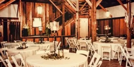 A rustic wooden event hall set up for a wedding with round tables covered in white linens, white Chiavari chairs, and floral centerpieces.