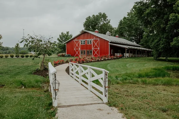 Exterior view of a large red barn with white trim and a white wooden bridge leading to the entrance, surrounded by green lawns and trees.