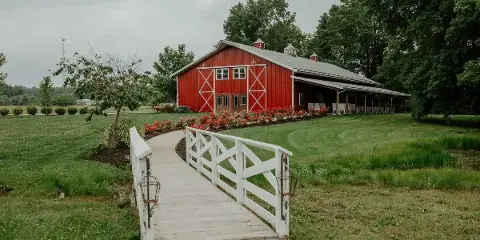 Exterior view of a large red barn with white trim and a white wooden bridge leading to the entrance, surrounded by green lawns and trees.