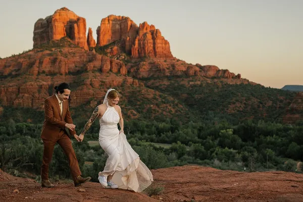 A bride and groom holding hands on a red rock ledge with Sedona's iconic buttes in the background.