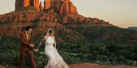 A bride and groom holding hands on a red rock ledge with Sedona's iconic buttes in the background.