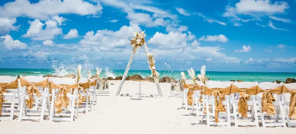 A beach wedding ceremony setup featuring a floral arch and rows of white chairs with gold sashes on the sand.
