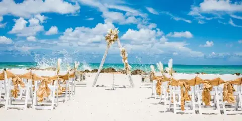 A beach wedding ceremony setup featuring a floral arch and rows of white chairs with gold sashes on the sand.