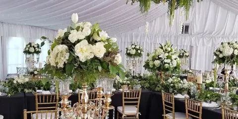 A wedding reception table decorated with large white and green floral arrangements, gold candelabras, and Chiavari chairs inside a white tent.