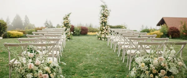 Rows of white cross-back chairs arranged on a lawn for a wedding ceremony with floral decorations.