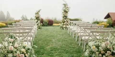 Rows of white cross-back chairs arranged on a lawn for a wedding ceremony with floral decorations.