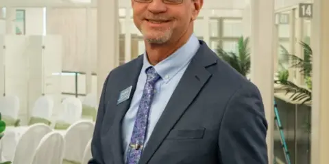 A man in a suit standing in front of a room with chairs decorated for a wedding ceremony.