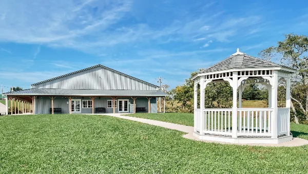 Exterior view of a grey metal barn building and a white wooden gazebo on a green lawn under a blue sky.