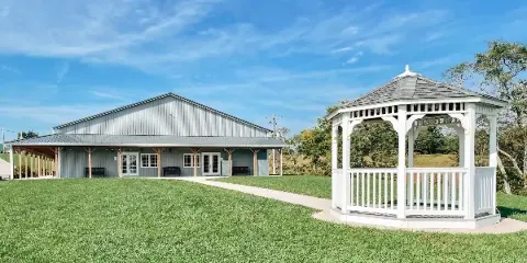 Exterior view of a grey metal barn building and a white wooden gazebo on a green lawn under a blue sky.