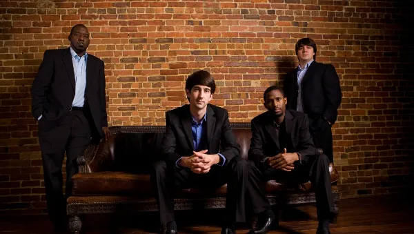 A group of four men in suits posing for a band portrait against a brick wall.