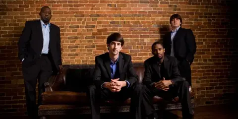 A group of four men in suits posing for a band portrait against a brick wall.