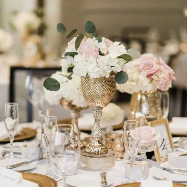 A close-up of a wedding table setting featuring a gold vase with white and pink flowers, glassware, and gold-rimmed plates.