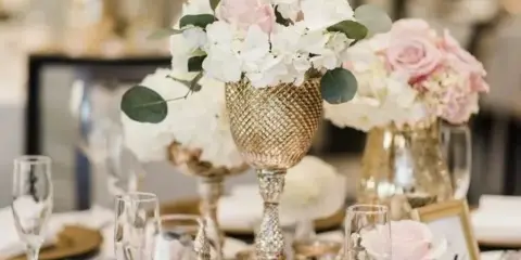A close-up of a wedding table setting featuring a gold vase with white and pink flowers, glassware, and gold-rimmed plates.
