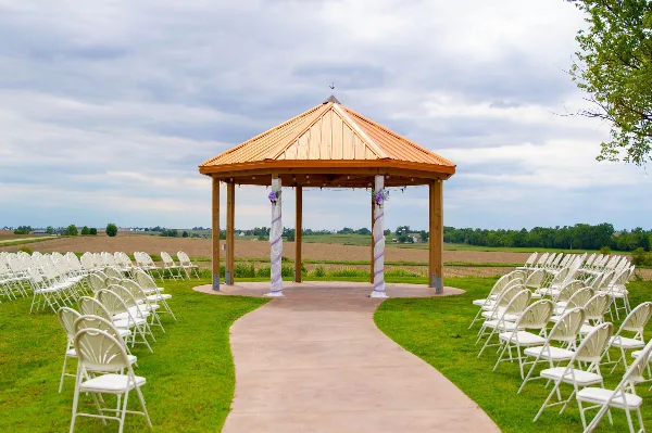 Outdoor wedding ceremony setup with white chairs arranged around a wooden gazebo in a grassy field.