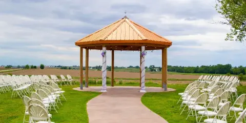 Outdoor wedding ceremony setup with white chairs arranged around a wooden gazebo in a grassy field.