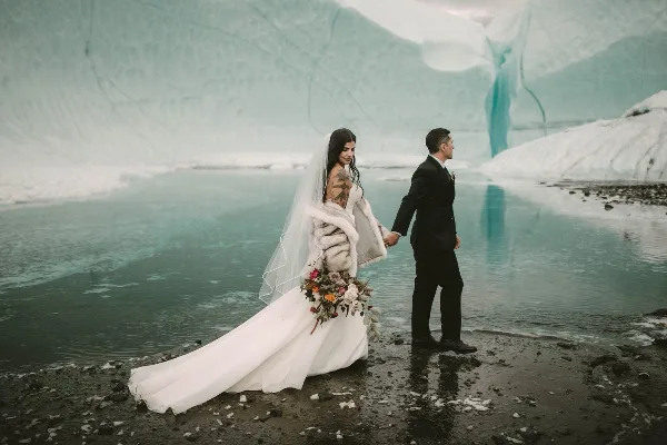 Bride and groom holding hands on a rocky shore in front of a blue glacier and meltwater pool.