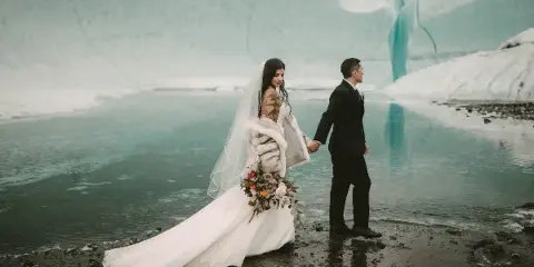 Bride and groom holding hands on a rocky shore in front of a blue glacier and meltwater pool.