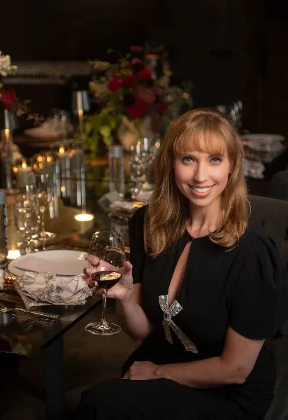 Portrait of a woman holding a glass of red wine, seated at a formally set event table with floral centerpieces and candles.
