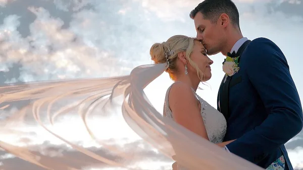 Bride and groom in a close embrace with the bride's veil flowing in the wind against a cloudy sky.