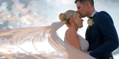 Bride and groom in a close embrace with the bride's veil flowing in the wind against a cloudy sky.