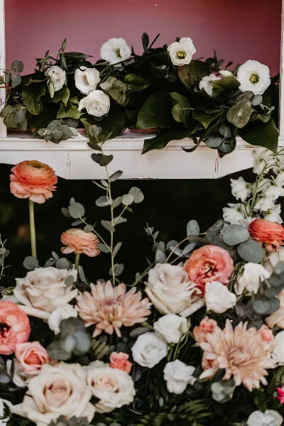 A white vintage-style wooden frame decorated with lush floral arrangements featuring white and peach roses, ranunculus, and eucalyptus.