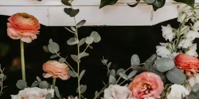 A white vintage-style wooden frame decorated with lush floral arrangements featuring white and peach roses, ranunculus, and eucalyptus.