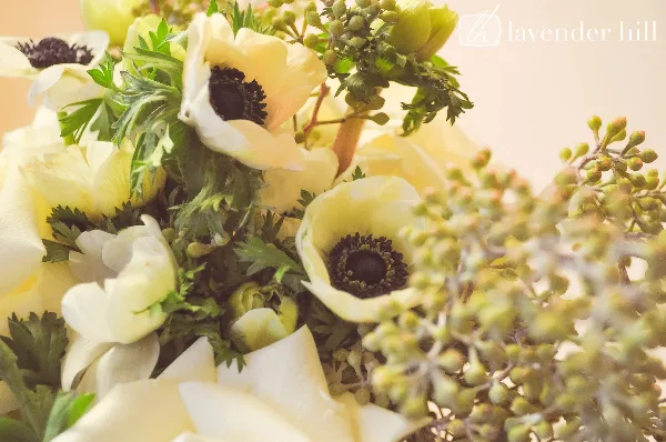 A close-up photograph of a floral arrangement featuring white anemones, cream roses, and green foliage.