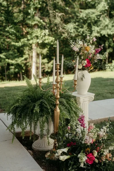 Outdoor event styling with a brass candelabra, ferns, and a floral arrangement in a white vase on a pedestal.