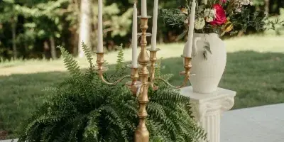 Outdoor event styling with a brass candelabra, ferns, and a floral arrangement in a white vase on a pedestal.