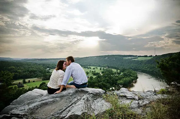 A couple seated on a rocky overlook with a river and green hills in the background.