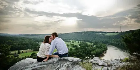 A couple seated on a rocky overlook with a river and green hills in the background.
