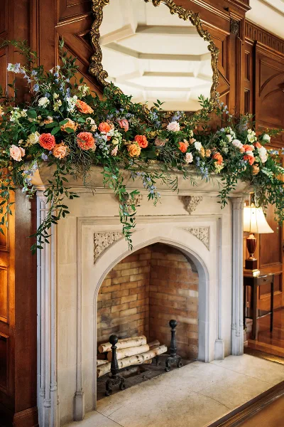 Floral arrangement with roses, greenery, and trailing vines decorating a stone fireplace mantel in a wood-paneled room.