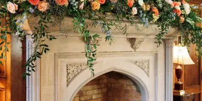 Floral arrangement with roses, greenery, and trailing vines decorating a stone fireplace mantel in a wood-paneled room.