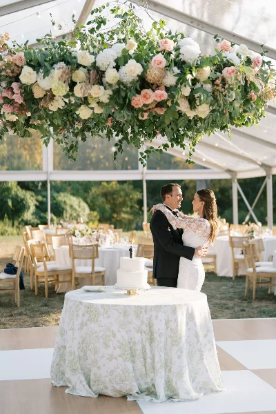 A bride and groom dancing at a wedding reception under a large floral installation in a white tent.