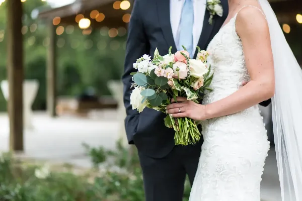 A bride in a white lace gown and a groom in a dark suit stand together holding a bouquet of white and pink flowers.