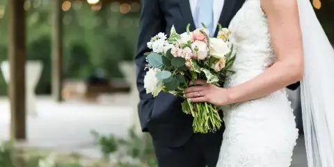 A bride in a white lace gown and a groom in a dark suit stand together holding a bouquet of white and pink flowers.