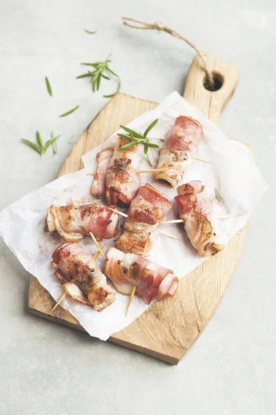 Bacon-wrapped appetizers served on a wooden board with parchment paper and rosemary garnish.