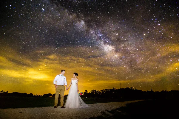 A bride and groom standing hand-in-hand under a starry night sky with the Milky Way visible.