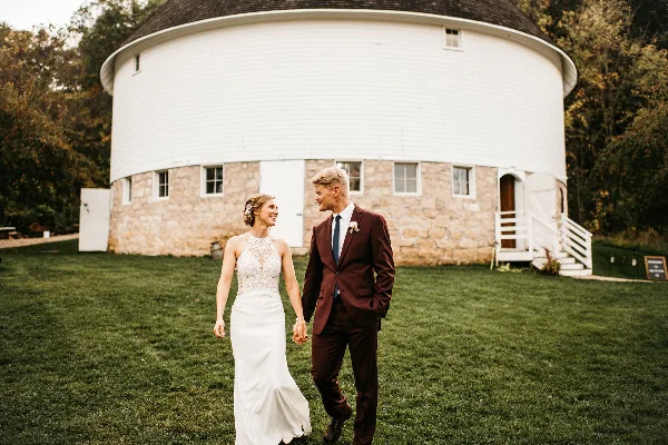 A bride and groom walking hand-in-hand on a lawn in front of a large white round barn with a stone foundation.