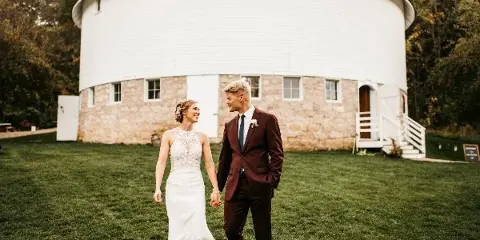 A bride and groom walking hand-in-hand on a lawn in front of a large white round barn with a stone foundation.