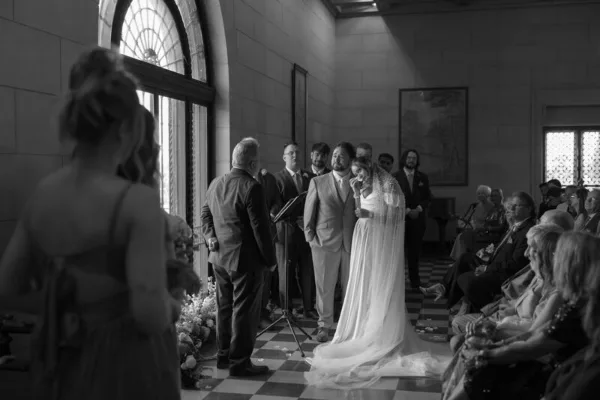 A black and white photograph of a bride and groom standing at the altar during a wedding ceremony in a stone-walled venue.