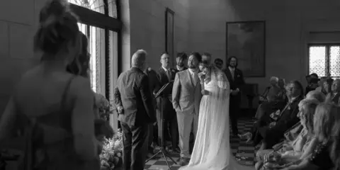 A black and white photograph of a bride and groom standing at the altar during a wedding ceremony in a stone-walled venue.
