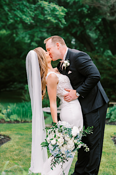 A bride in a white wedding dress holding a large bouquet of white and blue flowers while kissing a groom in a black suit.