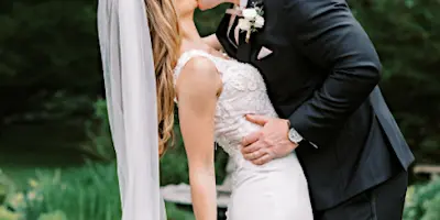 A bride in a white wedding dress holding a large bouquet of white and blue flowers while kissing a groom in a black suit.