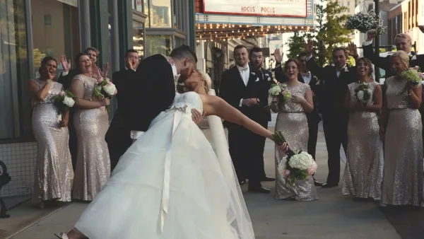 Cinematic wedding scene of a bride and groom kissing on a city street while the wedding party cheers and holds bouquets.